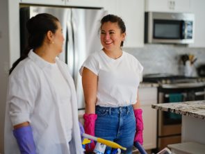 Dos mujeres con guantes de aseo conversan en la cocina de una casa