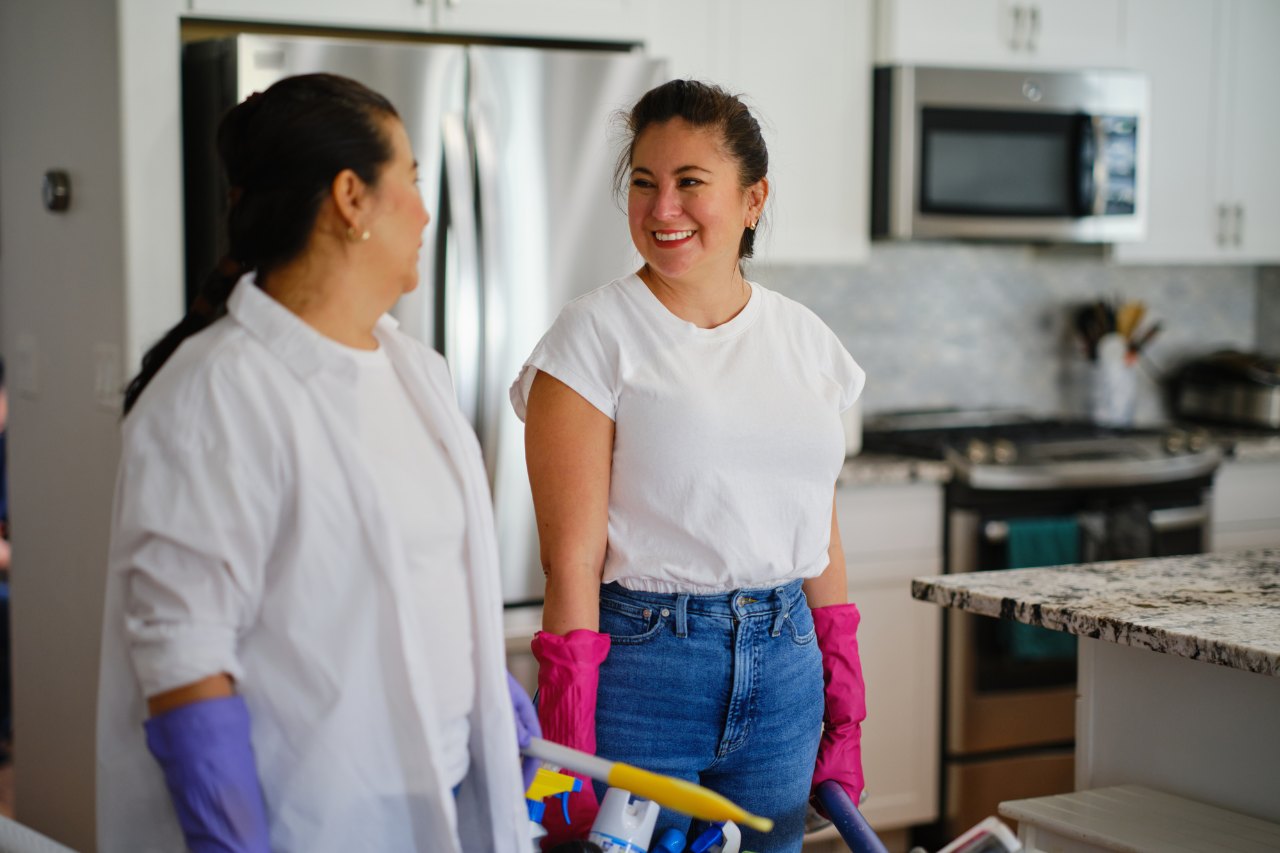 Dos mujeres con guantes de aseo conversan en la cocina de una casa