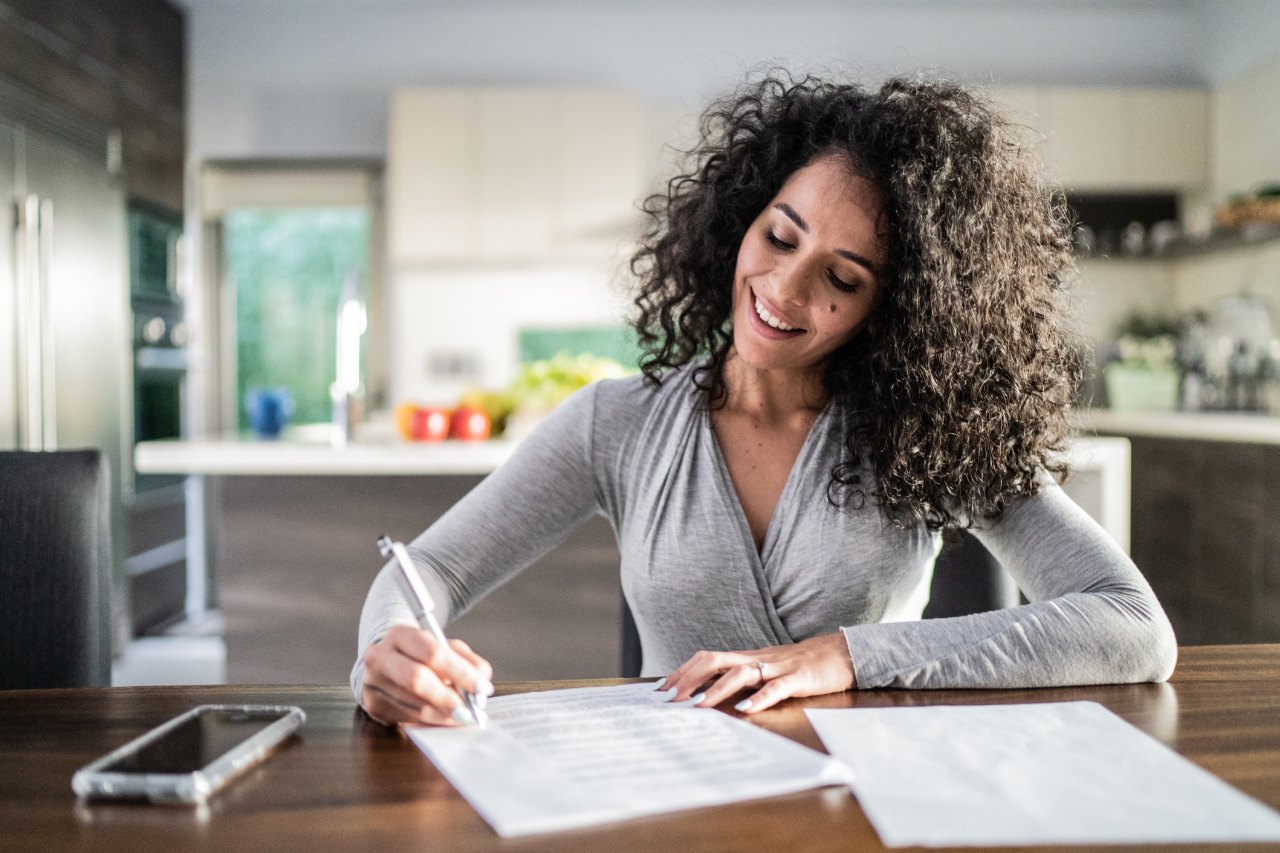 Mujer sonriente firma un documento sobre la mesa del comedor de su casa