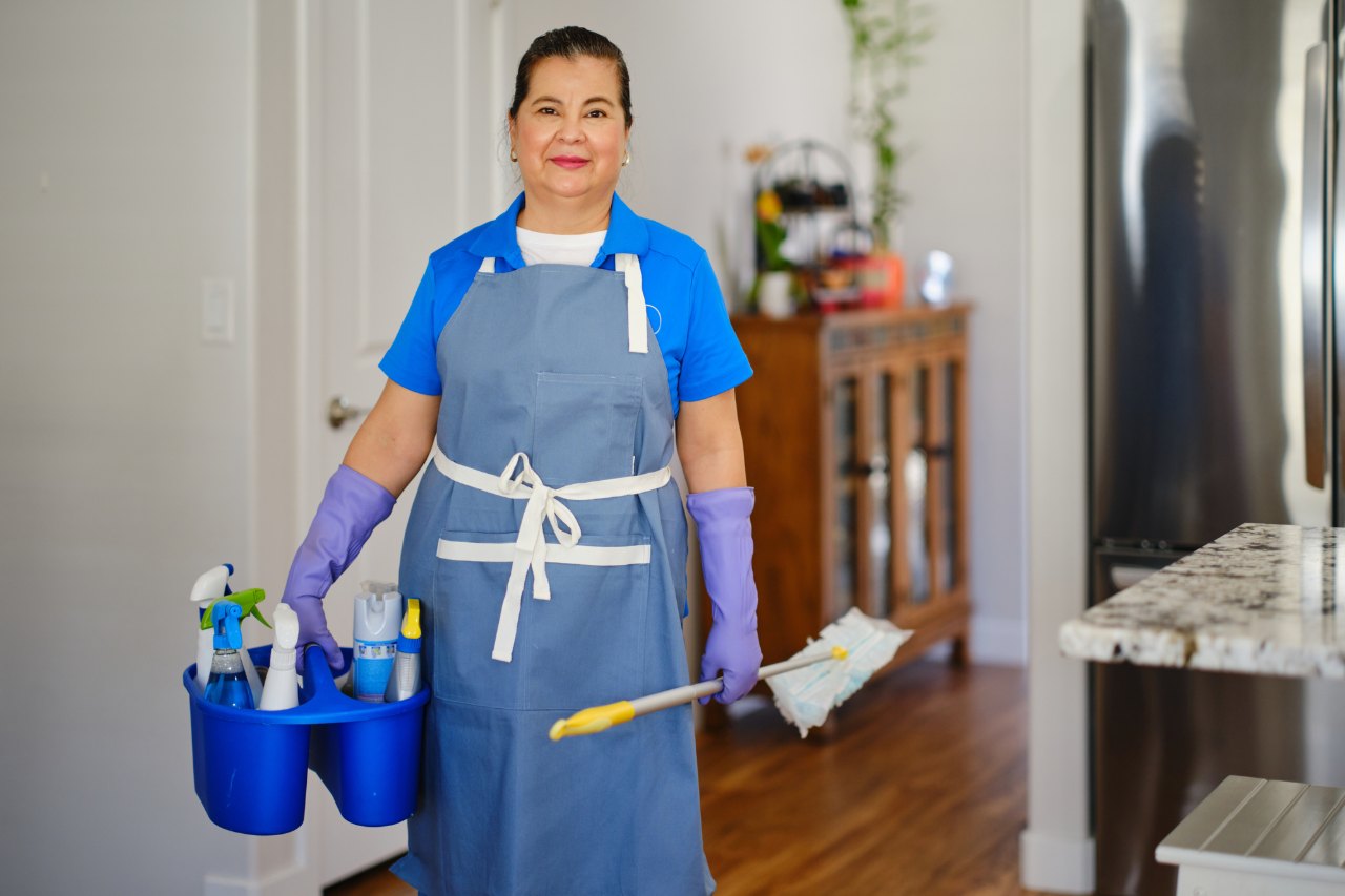 Empleada doméstica interna en 2026: salario, jornada, descansos y costo mensual 2 Empleada doméstica con uniforme azul, delantal y guantes sostiene implementos de aseo dentro de una casa