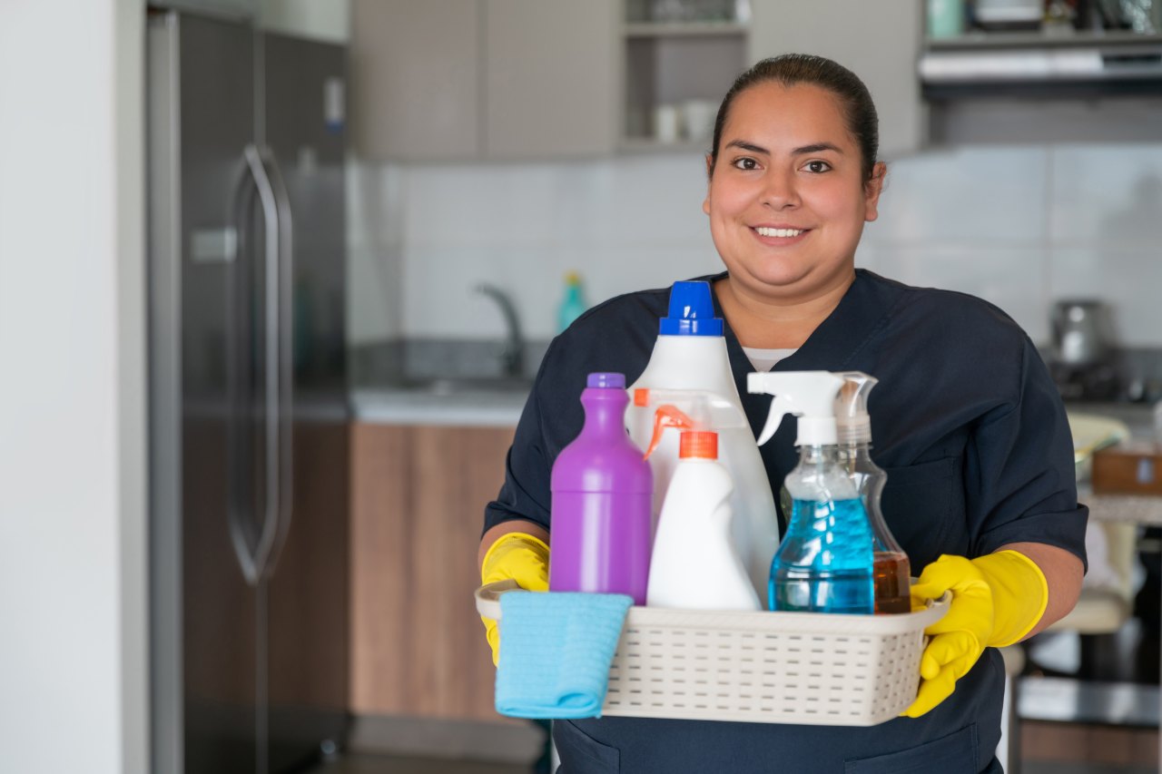 Empleada doméstica sonriente sostiene una canasta con implementos de aseo en una cocina