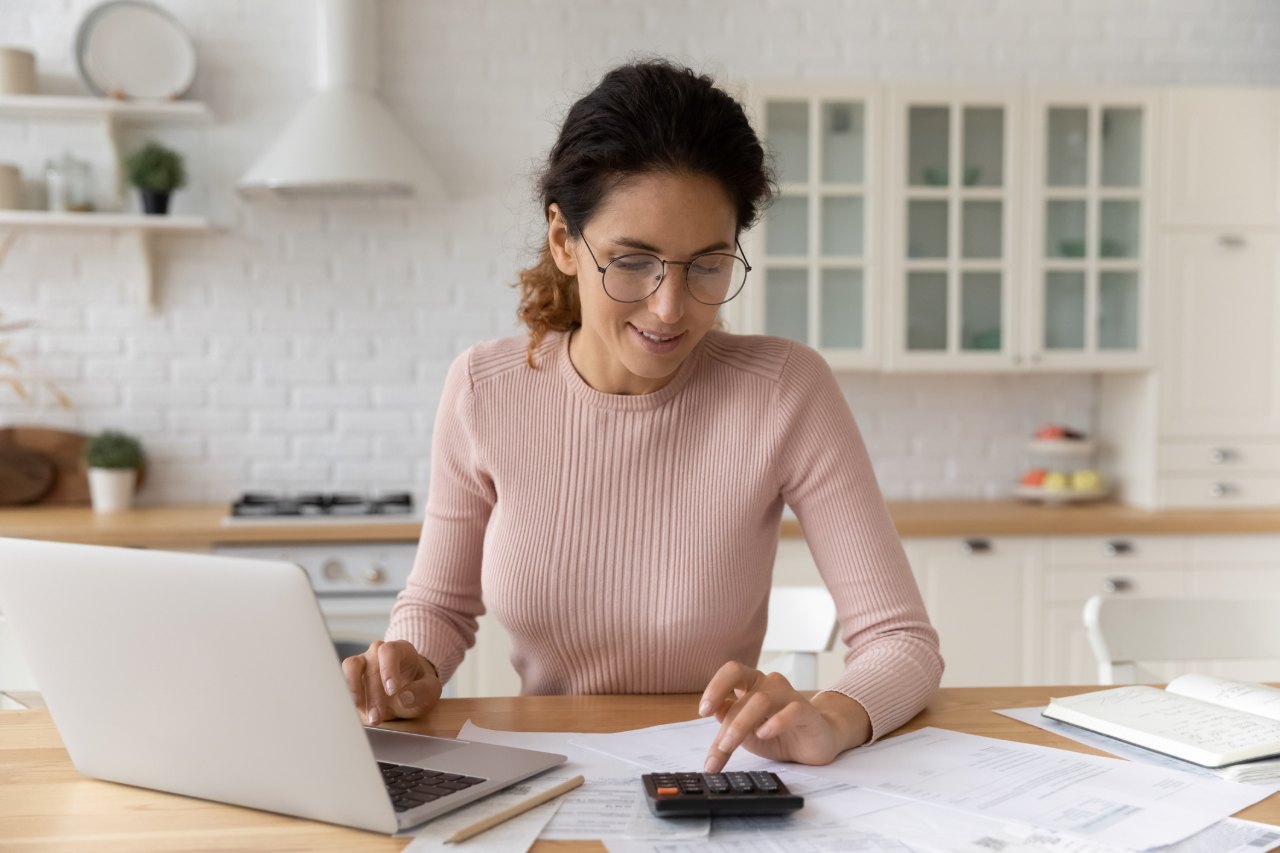 Mujer con gafas usa una calculadora frente a un computador portátil y documentos en la cocina de su casa