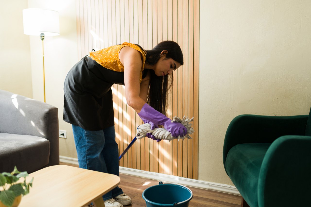 Señora de aseo con delantal y guantes escurre un trapero en la sala de una casa
