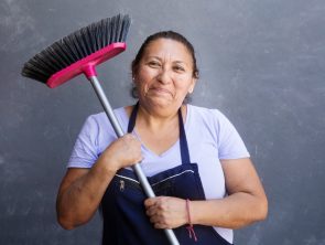Trabajadora doméstica con delantal sostiene una escoba sobre el hombro y sonríe