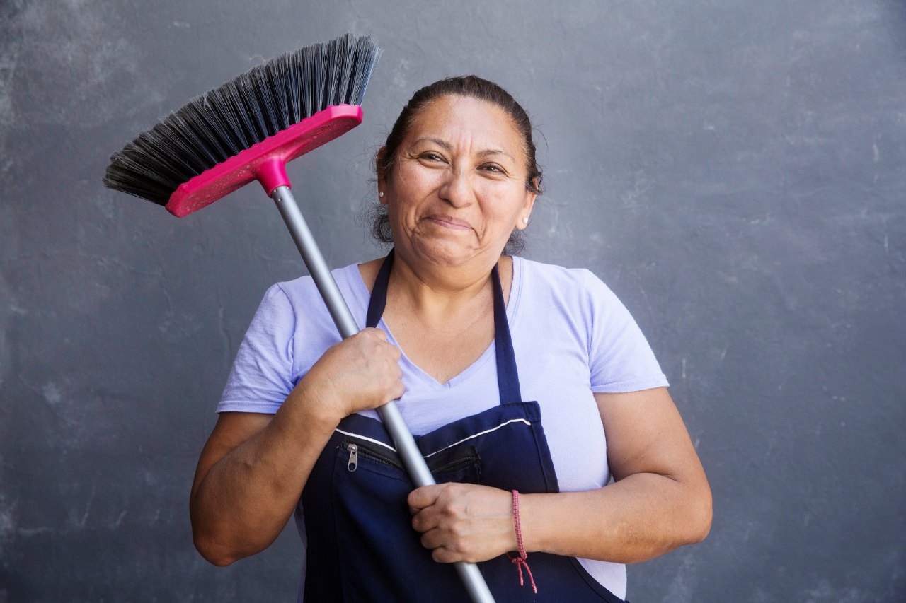 Trabajadora doméstica con delantal sostiene una escoba sobre el hombro y sonríe